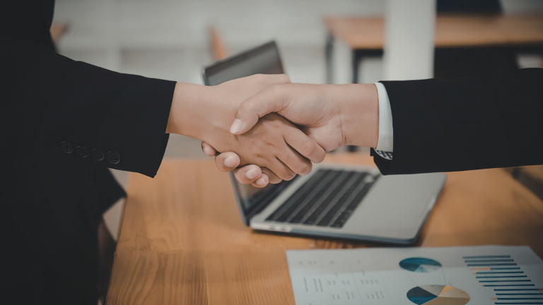 handshake between two people wearing suit jackets, you can only see the arms and hands. The background shows a wooden table, laptop and document with pie charts on it.