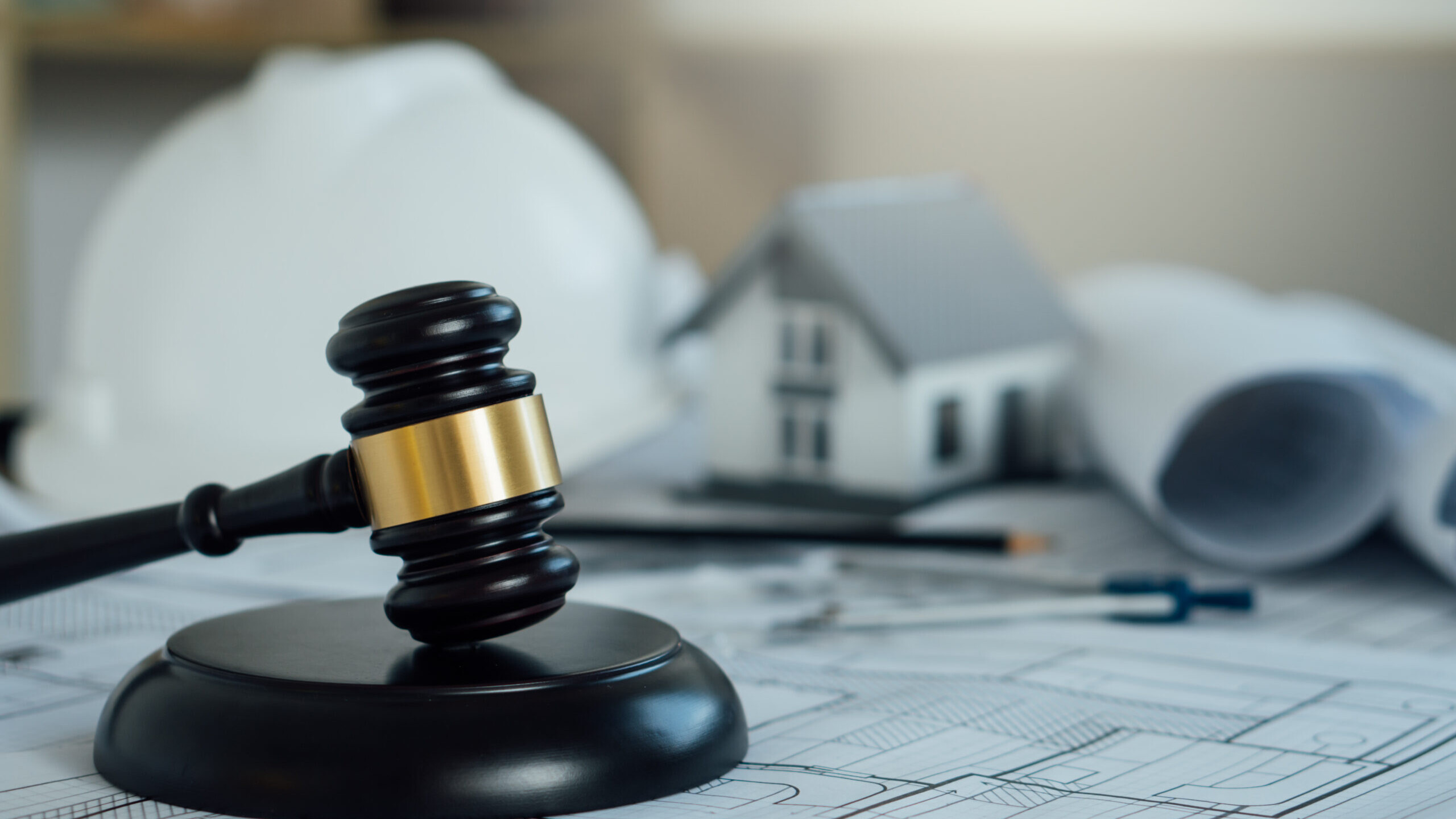 Gavel on architectural blueprints with a hard hat and model house in the background, representing real estate law and regulations.